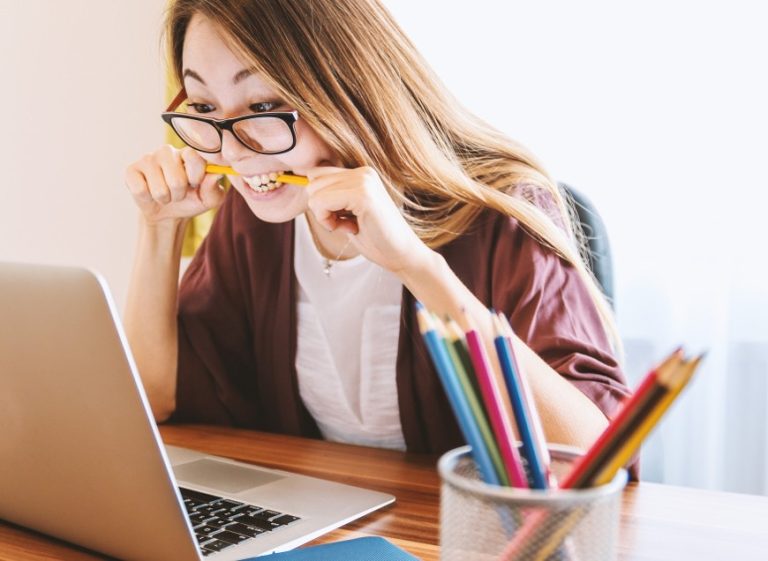 Stressed young woman biting a pencil while working on a laptop at a desk with colored pencils nearby.