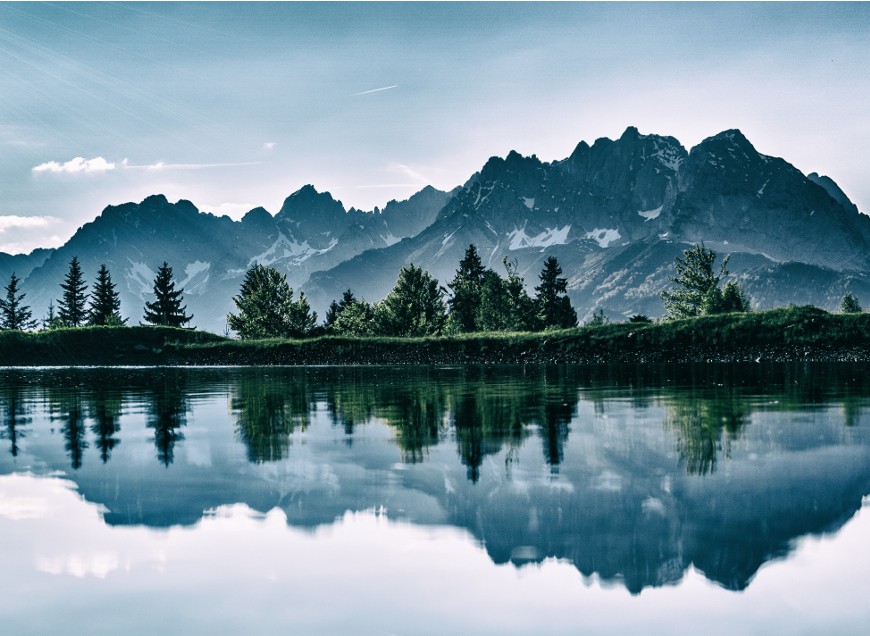 Snow-capped mountain range reflected in a calm lake with pine trees along the shoreline.