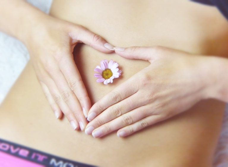 Hands forming a heart shape over a bare stomach with a small pink flower in the center.