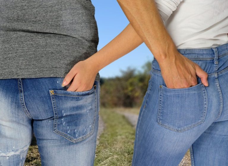 Two people standing side by side outdoors in blue jeans with their hands in their back pockets.